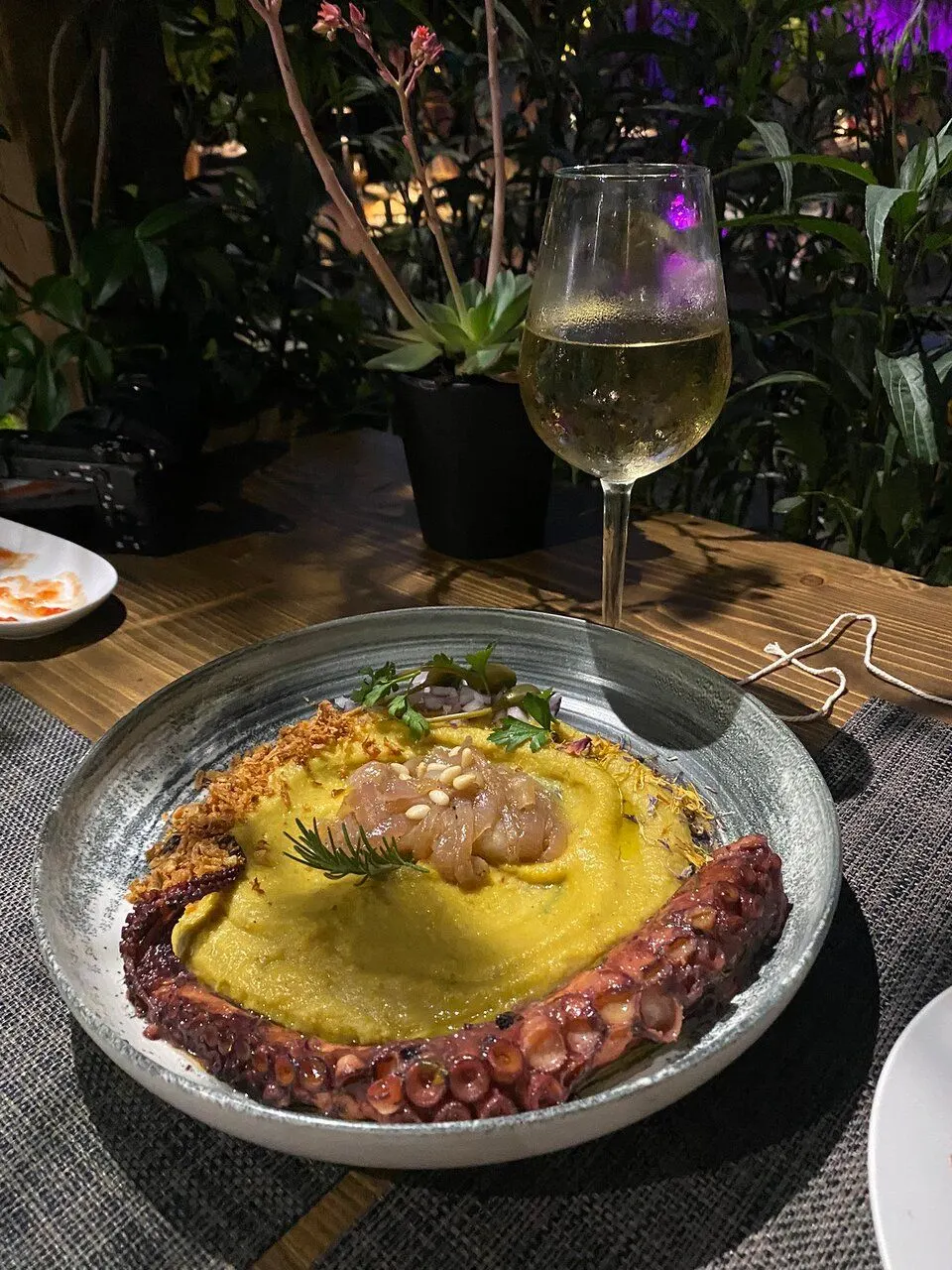 Grilled octopus curled over yellow fava purée in a stone bowl, topped with pickled onion and dill, photographed at the table at night with a glass of white wine behind it.