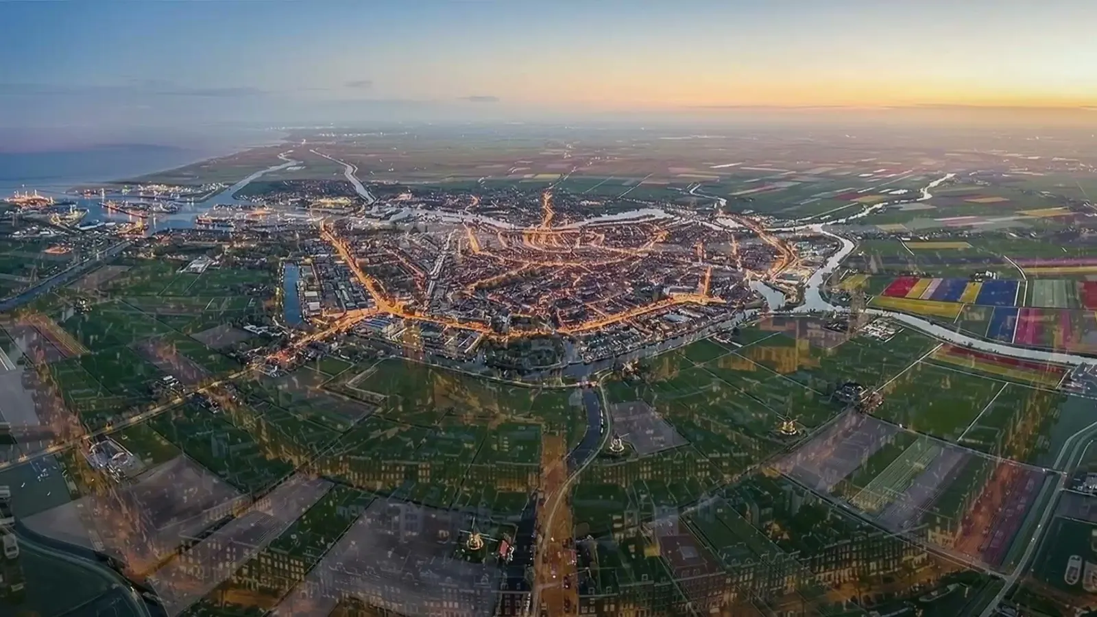 Amsterdam from high above at dusk, the canal ring lit in warm orange against blue water, the river and the coast visible at the top of the frame.