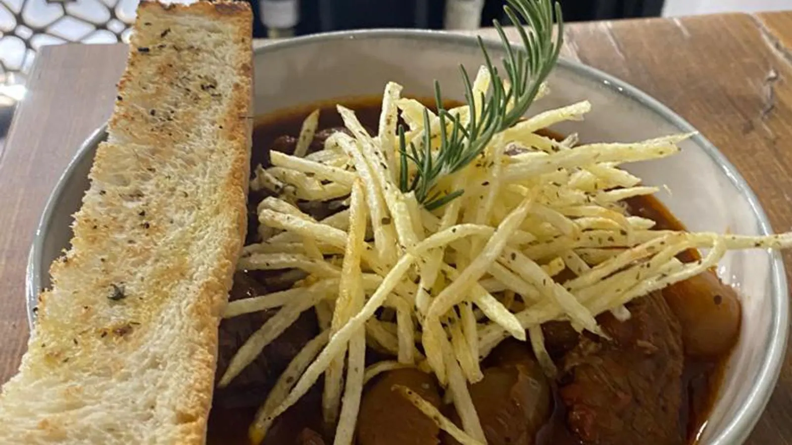 Beef stifado in a dark sauce in a metal bowl, under a nest of shoestring fries and a sprig of rosemary, a long slice of grilled bread beside it.