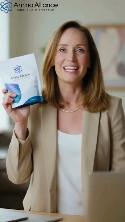A woman in a tan blazer holding an Amino Alliance pouch to camera in a bright office.
