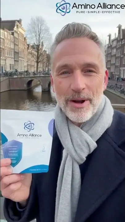 A man in a navy overcoat and grey scarf holding an Amino Alliance pouch to camera on an Amsterdam canal.