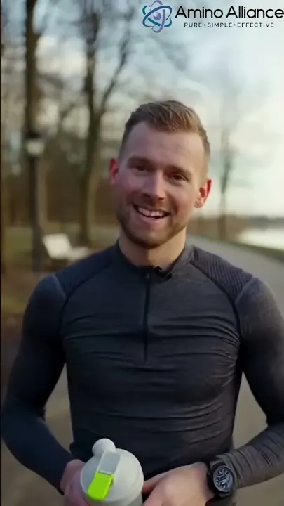 A man in a charcoal running top holding an Amino Alliance shaker on a park path in winter.