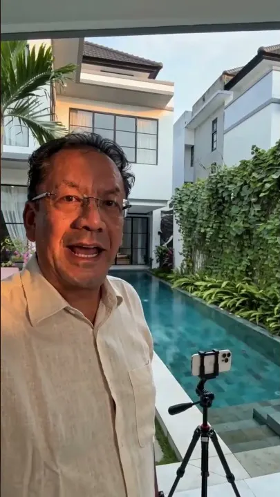 The presenter in a cream linen shirt speaking to camera beside a villa pool, a phone on a tripod set up facing him and planting up the wall behind.