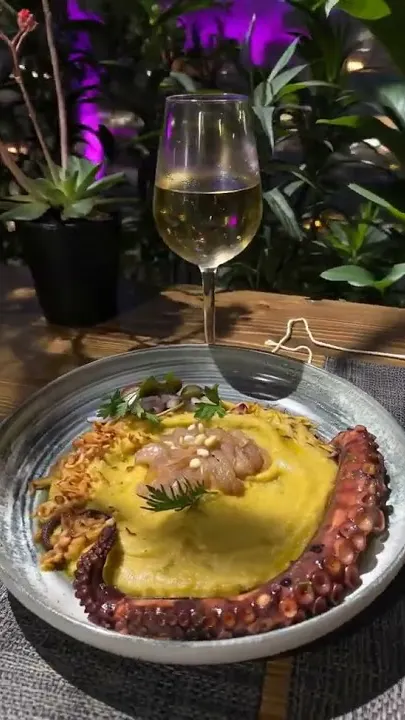 A frame from the 360 plate move: grilled octopus curled over yellow fava in a stone bowl on the terrace at night, a glass of white wine and lit planting behind it.